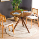 A solid oak round dining table with wooden butterfly legs, placed on a round white rug in a room with a gray wall and wooden floor.