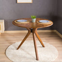 A solid oak round dining table with wooden butterfly legs, placed on a round white rug in a room with a gray wall and wooden floor.
