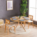 A solid oak round dining table with wooden butterfly legs, placed on a round white rug in a room with a gray wall and wooden floor.
